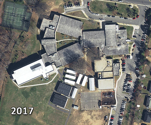 This series of aerial photographs shows Hybla Valley Elementary School from directly overhead in 2017, 2009, 2002, 1997, 1976, and 1937. In 2017, the two-story addition to our building is visible and several modular classrooms, trailers, and a basketball court can be seen behind the building. In 2009, the two-story wing and basketball court are gone, and there are more modular classrooms and trailers than in the 2017 photograph. In 2002, the playground has moved to a different location and there are fewer trailers behind the building. In 1997, the modular classrooms, most of the trailers, and the 8-classroom addition are gone. The playground is now on the west side of the building and cars can be seen parked on a basketball court that will later be turned into a parking lot. There are only 5 trailers outside the building. In 1976, the gymnasium and music room addition is gone. The building looks as it did when originally constructed, and is at least half the size of what it is today. The 1937 photograph is in black and white, and shows the school site when it was part of the airport property. Several buildings and trees are visible on what used to be farm fields.     
