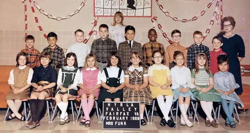 Color class photograph taken in February 1968. Pictured are students in Mrs. Funk’s class and she is standing on the far right of the picture in the background. 20 children are pictured, an even mix of boys and girls. The girls are seated in the front row and are all wearing dresses or skirts with knee socks. The boys stand behind them, all wearing button down shirts. Most of the shirts are plaid. One girl stands in the very back row. 