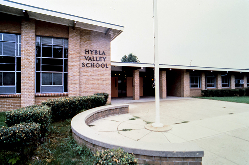 Color 35 millimeter slide photograph, taken in the early 1980s, showing the front of Hybla Valley Elementary School. The section pictured is the cafeteria, main entrance doors, and library. The cafeteria has since been remodeled and expanded outward. The doors are painted a bright orange. A flagpole is visible in the foreground, and the name of the school is on the side of the building in black letters. A window-mounted air conditioning unit is visible in one of the library windows.    